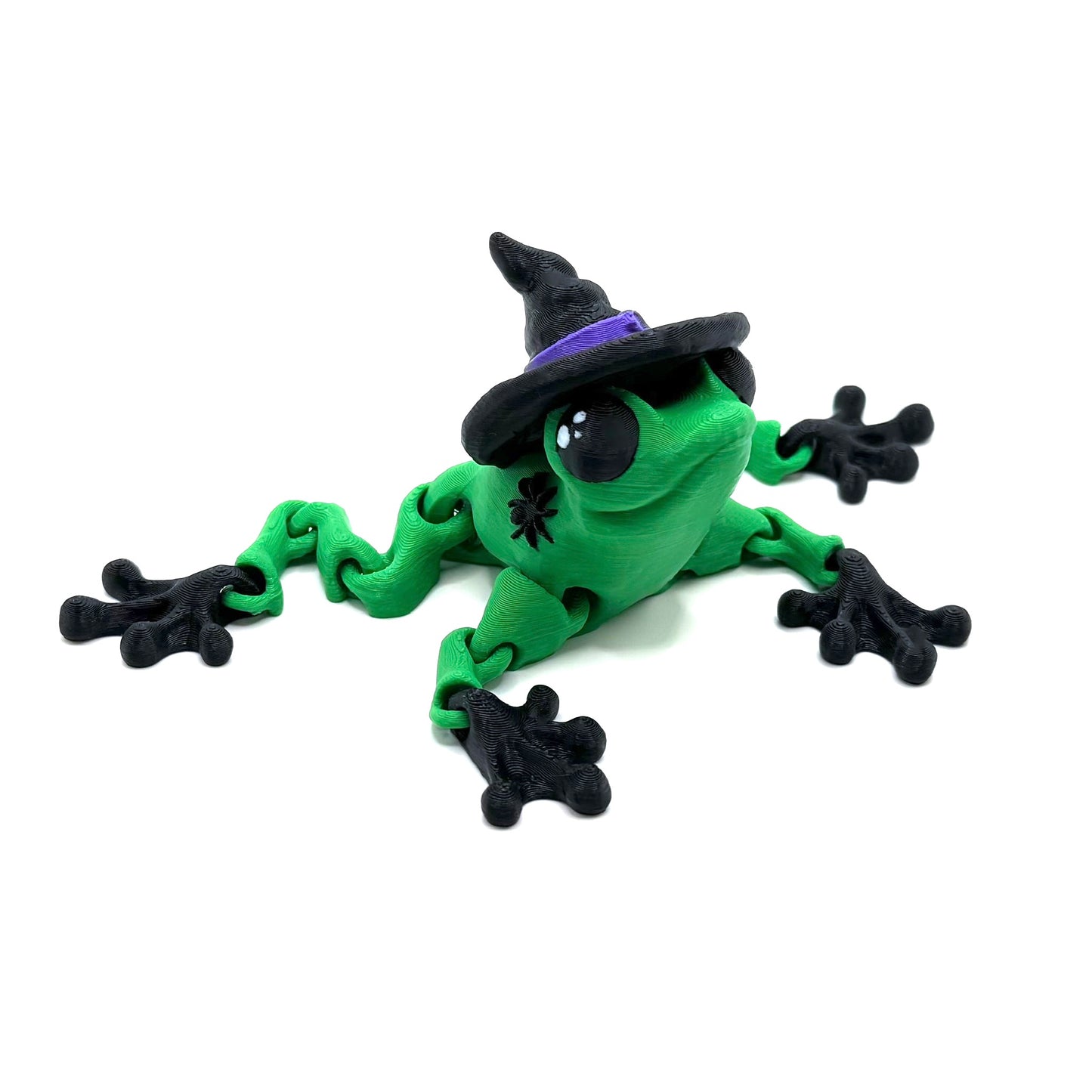 A green witch frog with jointed limbs wearing a black witch hat with a purple band, sitting on a white surface.
