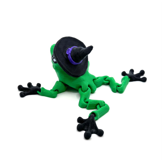 Back view of a green frog with black feet, wearing a black witch hat with a purple band, sitting on a white background.