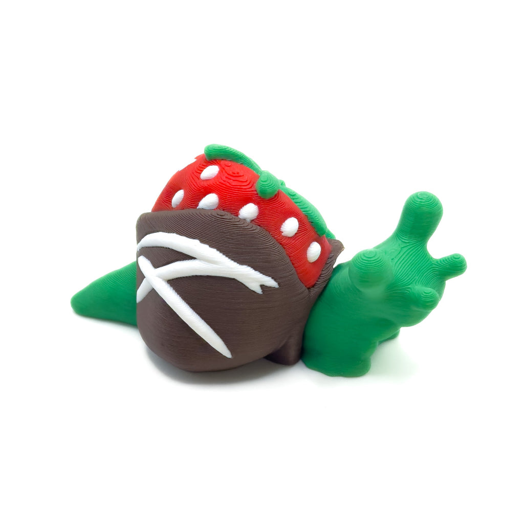 A green snail with a red strawberry shell dipped in chocolate and decorated with white stripes, resting on a white background.